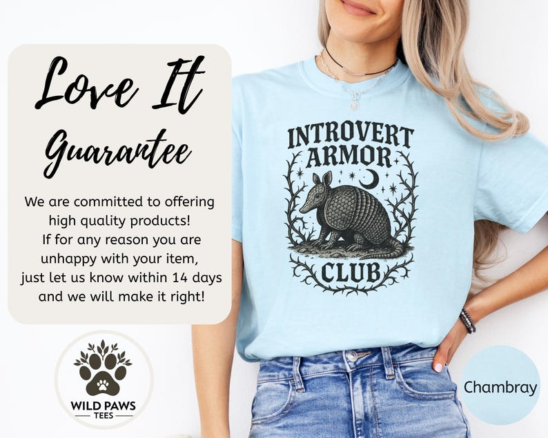 Op de afbeelding: Lichtblauw T-shirt met de tekst "INTROVERT ARMOR CLUB" en een gedetailleerde illustratie van een gordeldier. Het shirt heeft ook het "WILD PAWS TEES" logo en het woord "Chambray".