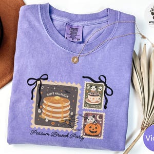 Puede incluir: Una camiseta violeta Comfort Colors con un diseño de Halloween. El diseño incluye ilustraciones de sellos postales de panqueques, una zarigüeya en una taza de té y una calabaza. También se ve el texto "Happy Halloween" y "Possum Brunch Party".