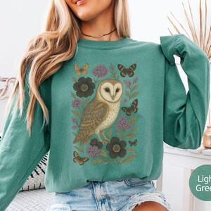 Puede incluir: Sudadera de manga larga verde claro con una ilustraci&oacute;n detallada de un b&uacute;ho com&uacute;n, mariposas y flores. El b&uacute;ho es marr&oacute;n y blanco, las mariposas son marrones y las flores son moradas y negras.