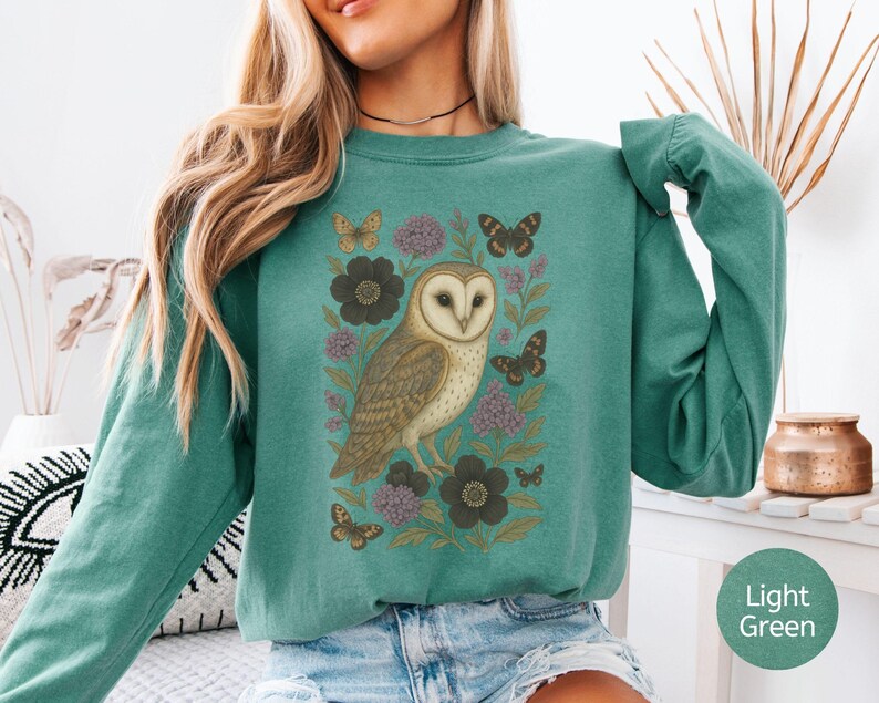 Puede incluir: Sudadera de manga larga verde claro con una ilustraci&oacute;n detallada de un b&uacute;ho com&uacute;n, mariposas y flores. El b&uacute;ho es marr&oacute;n y blanco, las mariposas son marrones y las flores son moradas y negras.
