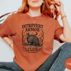 Op de afbeelding: Een roestkleurig t-shirt met de tekst "Introvert Armor Club" boven een gedetailleerde illustratie van een gordeldier, een halve maan en sterren. Het ontwerp is omlijst door een decoratieve rand. Het shirt is effen van kleur.