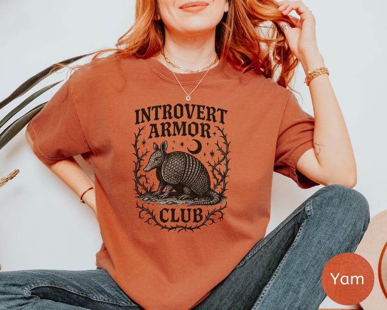 Op de afbeelding: Een roestkleurig t-shirt met de tekst "Introvert Armor Club" boven een gedetailleerde illustratie van een gordeldier, een halve maan en sterren. Het ontwerp is omlijst door een decoratieve rand. Het shirt is effen van kleur.