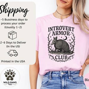 Op de afbeelding: Lichtroze t-shirt met de tekst "INTROVERT ARMOR CLUB" en een gedetailleerde illustratie van een gordeldier. Het shirt is gemaakt van een zacht materiaal en is geschikt voor vrijetijdskleding.