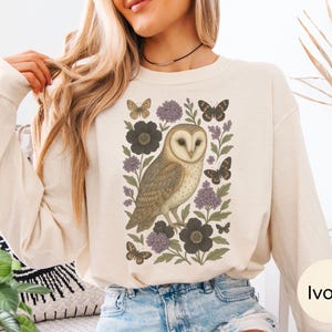 Puede incluir: Sudadera de manga larga color marfil con una ilustraci&oacute;n detallada de un b&uacute;ho rodeado de mariposas y elementos florales en tonos marr&oacute;n, morado y negro. El dise&ntilde;o est&aacute; centrado en la parte delantera.