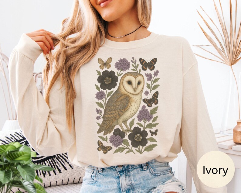 Puede incluir: Sudadera de manga larga color marfil con una ilustraci&oacute;n detallada de un b&uacute;ho rodeado de mariposas y elementos florales en tonos marr&oacute;n, morado y negro. El dise&ntilde;o est&aacute; centrado en la parte delantera.
