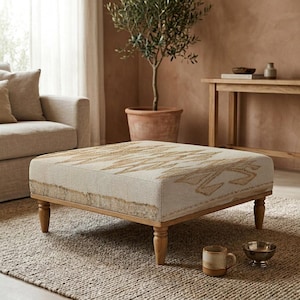 May include: A square ottoman with a light beige fabric top featuring a geometric pattern in shades of tan and gold. The ottoman has a light wood frame and four rounded legs. The room has a neutral color palette.