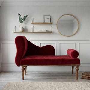 May include: A deep red velvet chaise lounge featuring a high, curved backrest on one side and a cylindrical bolster pillow on the other. The piece is supported by four turned wooden legs with a natural finish, showcasing a smooth, plush fabric texture.
