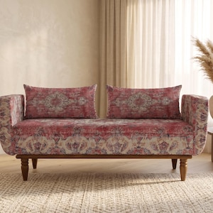 Op de afbeelding: Een tweezitsbank met een rood en beige patroon. De bank heeft een houten frame en poten. Twee bijpassende rechthoekige kussens liggen op de bank. De bank staat in een kamer met een vloerkleed en een bijzettafel.