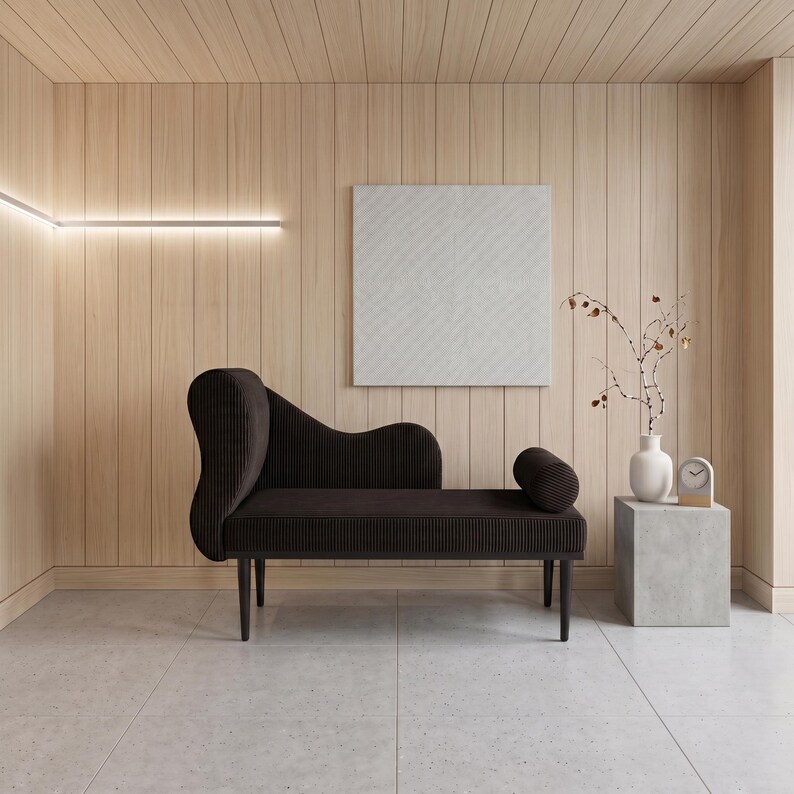 May include: A modern interior scene featuring a dark brown chaise lounge with a curved back and cylindrical bolster. The room has light wood paneling, a white square artwork, and a small side table with a vase and clock.