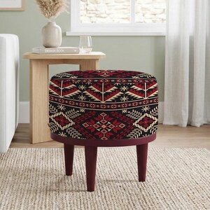 May include: A round ottoman with a patterned top featuring red, black, and beige geometric designs. The ottoman has a burgundy-colored base and legs, and is placed on a neutral-colored rug.