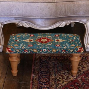 Peut inclure: Un petit pouf rectangulaire avec un motif floral turquoise, rouge et crème. Le pouf a quatre pieds en bois et est placé sous une grande chaise rembourrée avec des pieds blancs ornés. Un tapis à motifs est visible.