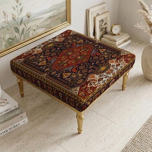 May include: A square ottoman with a patterned top in shades of red, blue, and gold, set on four gold-coloured legs. The ottoman is placed in a room with a neutral colour palette, with a vase of dried flowers and a stack of books nearby.