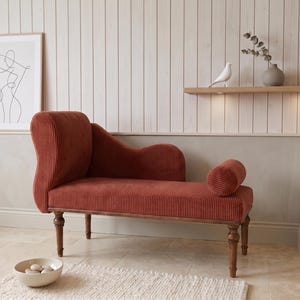 May include: A rust-colored chaise lounge with a corduroy texture, wooden legs, and a rolled bolster pillow. The elegant furniture piece is set against a neutral-toned wall with a framed art print and a decorative shelf with a bird figurine.