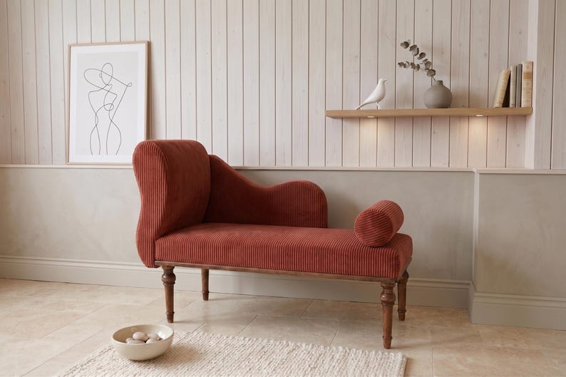 May include: A rust-colored chaise lounge with a corduroy texture, wooden legs, and a rolled bolster pillow. The elegant furniture piece is set against a neutral-toned wall with a framed art print and a decorative shelf with a bird figurine.