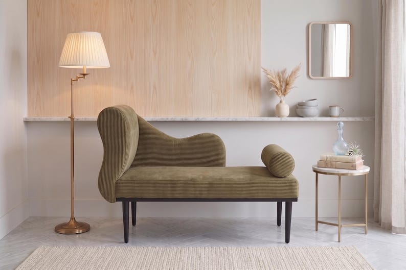 K&ouml;nnte beinhalten: Eine olivgr&uuml;ne Chaiselongue mit geschwungener R&uuml;ckenlehne und Kissenrolle. Eine Messing-Stehlampe mit plissiertem Lampenschirm steht links. Ein kleiner Beistelltisch mit Marmorplatte und ein dekorativer Spiegel befinden sich ebenfalls im Raum.