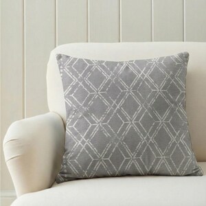 Puede incluir: Un cojín decorativo cuadrado con un patrón geométrico de diamantes en gris y blanco. El cojín está colocado sobre un sofá blanco, ofreciendo una paleta de colores neutros para la decoración del hogar.