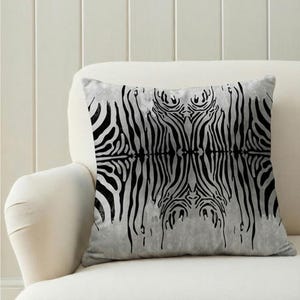 May include: A square cushion featuring a black and white zebra stripe design. The pattern is mirrored, producing a symmetrical image. The cushion is placed on a white sofa.