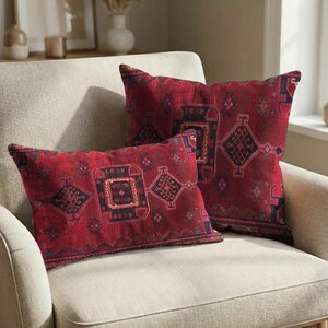May include: Two decorative throw pillows with a red background and geometric patterns in navy, brown, and beige. The pillows are placed on a beige armchair, creating a cozy and inviting atmosphere.