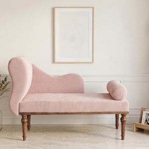 May include: A pink corduroy chaise lounge with a curved back and a cylindrical bolster. The wooden legs and frame support the seating. A framed abstract art piece hangs on the wall behind the chaise lounge.