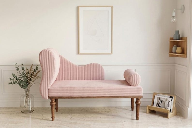 May include: A pink corduroy chaise lounge with a curved back and a cylindrical bolster. The wooden legs and frame support the seating. A framed abstract art piece hangs on the wall behind the chaise lounge.
