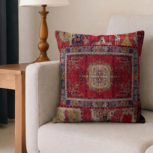 May include: A decorative throw pillow with a red, patterned design reminiscent of a traditional rug. The pillow features a central medallion and intricate border details in shades of blue, yellow, and cream. It rests on a neutral-toned sofa.