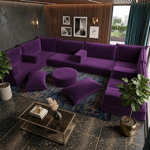 May include: A large U-shaped purple velvet sectional sofa with matching rectangular and round floor cushions. The set includes a low-profile modular design with backrests and armrests, arranged on a dark blue patterned rug in a room.
