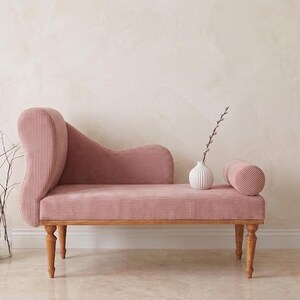 May include: A pink corduroy chaise lounge with wooden legs and a curved back. A small white vase with a sprig of greenery and a cylindrical bolster pillow sit on the seat. Decorative vases with dried flowers are on the floor.