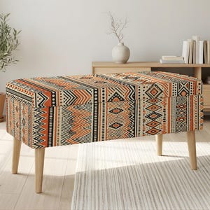 May include: A rectangular storage bench with a geometric tribal pattern in orange, black, and cream. The bench has four wooden legs and is placed on a light-colored rug. The bench is a decorative and functional piece of furniture.