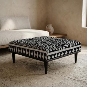 Peut inclure: Un pouf carré avec un dessus à motifs noir et blanc et un cadre en bois noir. Le dessus présente un motif floral détaillé. Le pouf a quatre pieds noirs et est placé sur un sol en pierre. Le design est un mélange de styles modernes et bohèmes.