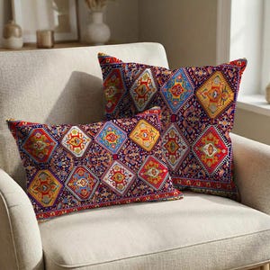 Op de afbeelding: Twee decoratieve sierkussens met een levendig, geometrisch patroon in tinten rood, blauw, geel en oranje op een donkerpaarse achtergrond. De kussens zijn rechthoekig en vierkant, geplaatst op een beige fauteuil.
