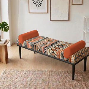 May include: A modern bench with a black frame, featuring a patterned seat in orange, black, and cream geometric designs. It has two orange bolster pillows and is set on a wooden floor.