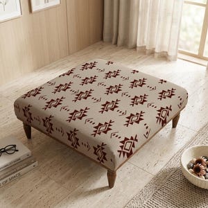 Peut inclure: Un pouf carré avec un tissu beige orné d'un motif géométrique répétitif rouge foncé. Le pouf a des pieds en bois et est placé sur un sol carrelé. Une pile de livres et un bol de pierres décoratives sont à proximité.