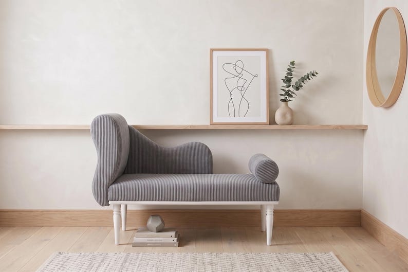 May include: A gray chaise lounge with a ribbed texture, white legs, and a curved back. The furniture is in a room with a framed art print, a vase with greenery, and a round wooden mirror. The room has a neutral color palette.