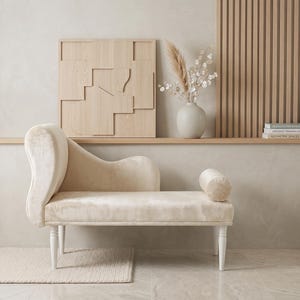May include: A cream-colored velvet chaise lounge featuring a curved backrest, a single cylindrical bolster pillow, and four white tapered legs. The upholstery has a soft, plush texture, and the frame design includes a high, rounded armrest.