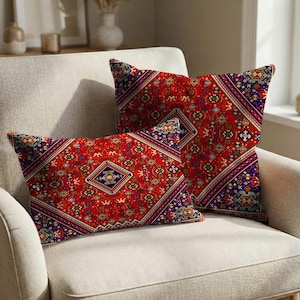 Op de afbeelding: Twee decoratieve kussens met een levendig rood, blauw en goud geometrisch patroon. De kussens zijn rechthoekig en vierkant, geplaatst op een neutrale fauteuil. Het ontwerp lijkt op een traditioneel tapijt.
