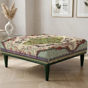 Pode incluir: Um pufe retangular com um tampo de tecido estampado com um design floral verde, vermelho e creme. O pufe tem uma estrutura e pernas de madeira verde escuro e está colocado numa sala com paredes e pisos de cores claras.