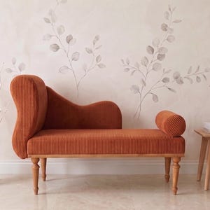 May include: An orange corduroy chaise lounge with a curved back and armrest. The furniture piece has wooden legs and is placed in front of a wall with a floral design. A vase with silver branches and a small side table with a vase and books are also present.