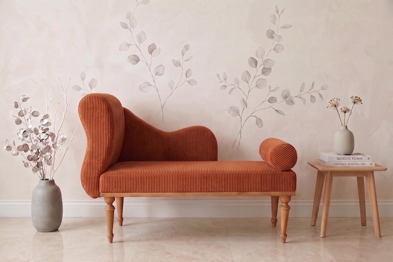 May include: An orange corduroy chaise lounge with a curved back and armrest. The furniture piece has wooden legs and is placed in front of a wall with a floral design. A vase with silver branches and a small side table with a vase and books are also present.