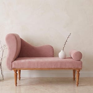 May include: A pink corduroy chaise lounge with wooden legs and a curved back. A small white vase with a sprig of greenery and a cylindrical bolster pillow sit on the seat. Decorative vases with dried flowers are on the floor.