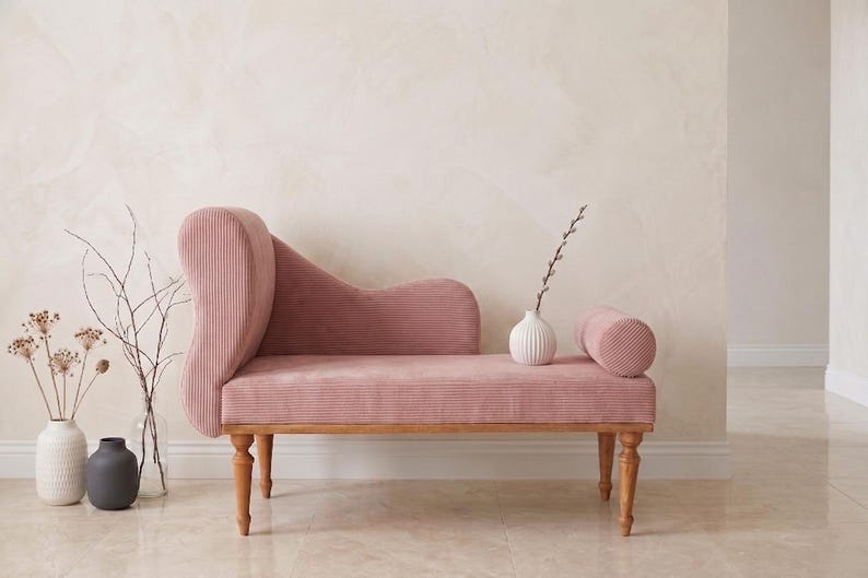 May include: A pink corduroy chaise lounge with wooden legs and a curved back. A small white vase with a sprig of greenery and a cylindrical bolster pillow sit on the seat. Decorative vases with dried flowers are on the floor.