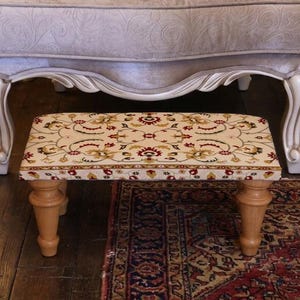 May include: A small, rectangular footstool with a patterned top and wooden legs. The fabric features a cream background with a repeating floral design in red, yellow, and green. The legs are a light brown color.