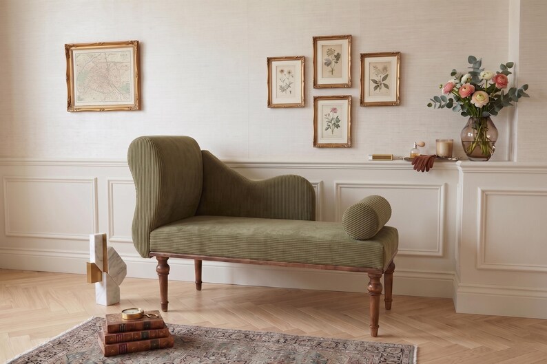 May include: An olive green chaise lounge with a ribbed texture, wooden legs, and a rolled bolster. The room features framed botanical prints, a map, and a vase of flowers. The decor is complemented by a patterned rug and wooden flooring.