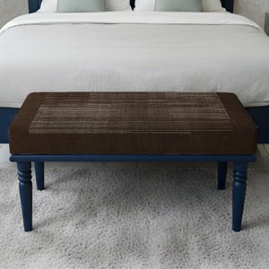 May include: A brown rectangular bench with a patterned top and dark blue legs sits at the foot of a bed. The bed has a dark blue frame and a white duvet. The bench is on a light grey rug. The bench is approximately 120 cm long.