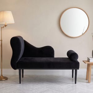 May include: A black velvet chaise lounge with a curved backrest and a cylindrical bolster pillow. The furniture piece features a solid black frame supported by four tapered black legs, providing a sleek and low-profile seating arrangement.