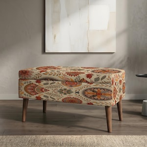 Puede incluir: Otomana tapizada con un estampado floral en tonos naranja, rojo y marrón sobre un fondo crema. La otomana tiene cuatro patas de madera y está colocada sobre un suelo de madera. La habitación tiene una paleta de colores neutros con una ventana grande.