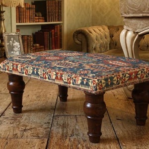 Puede incluir: Un otomán rectangular con una parte superior de tela estampada en tonos azules, rojos y crema. Tiene cuatro patas de madera torneada de color marrón oscuro. El otomán está sobre un suelo de madera, típico de un salón.