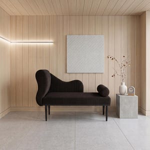 May include: A modern interior scene featuring a dark brown chaise lounge with a curved back and cylindrical armrest. The room has light wood paneling, a white square artwork, and a small side table with a vase and clock.