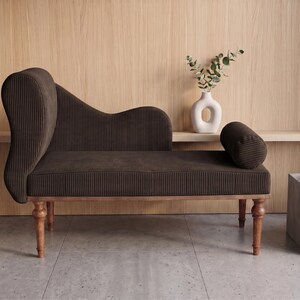 K&ouml;nnte beinhalten: Eine braune Chaiselongue mit hohem R&uuml;cken und einem zylindrischen Kissen. Die M&ouml;bel haben einen Holzrahmen und Beine. Eine wei&szlig;e Vase mit Gr&uuml;n und ein kleiner Beistelltisch mit einem goldenen Tablett sind ebenfalls zu sehen.