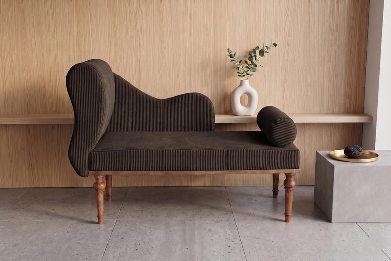 K&ouml;nnte beinhalten: Eine braune Chaiselongue mit hohem R&uuml;cken und einem zylindrischen Kissen. Die M&ouml;bel haben einen Holzrahmen und Beine. Eine wei&szlig;e Vase mit Gr&uuml;n und ein kleiner Beistelltisch mit einem goldenen Tablett sind ebenfalls zu sehen.