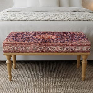 May include: A rectangular bench with a patterned top and wooden legs. The top features a traditional rug design in shades of red, blue, and cream. The bench is placed at the foot of a bed with a neutral-coloured bedspread.